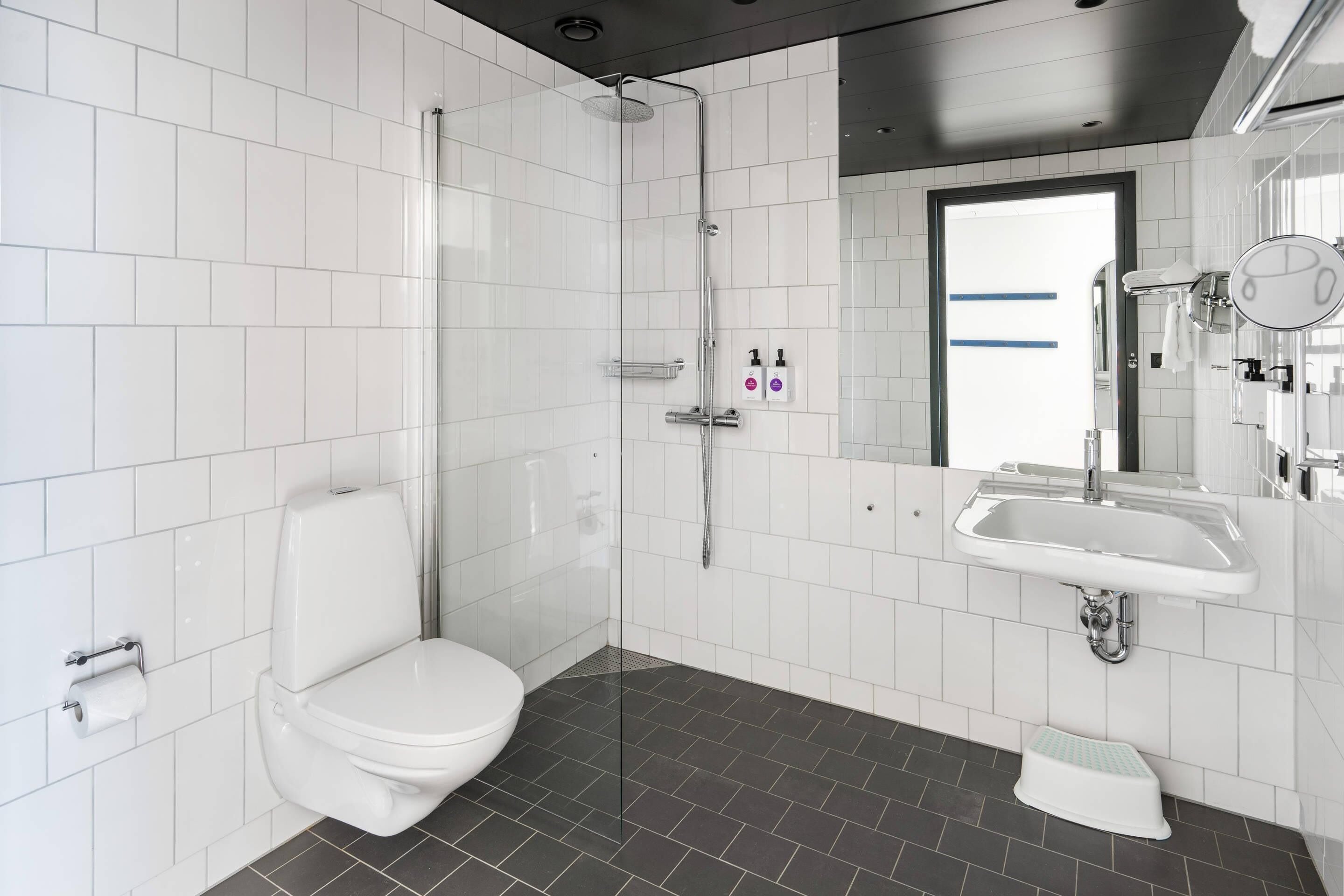 Ein modernes Badezimmer mit weißen Fliesen, einer Dusche, einer Toilette, einem Waschbecken und einem Spiegel.