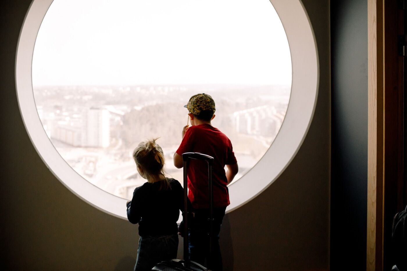 Two children looking out from a round window with a view of a cityscape.