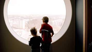 Two children looking out from a round window with a view of a cityscape.