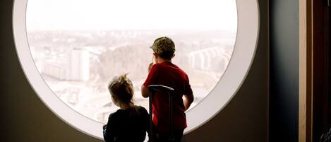Two children looking out from a round window with a view of a cityscape.
