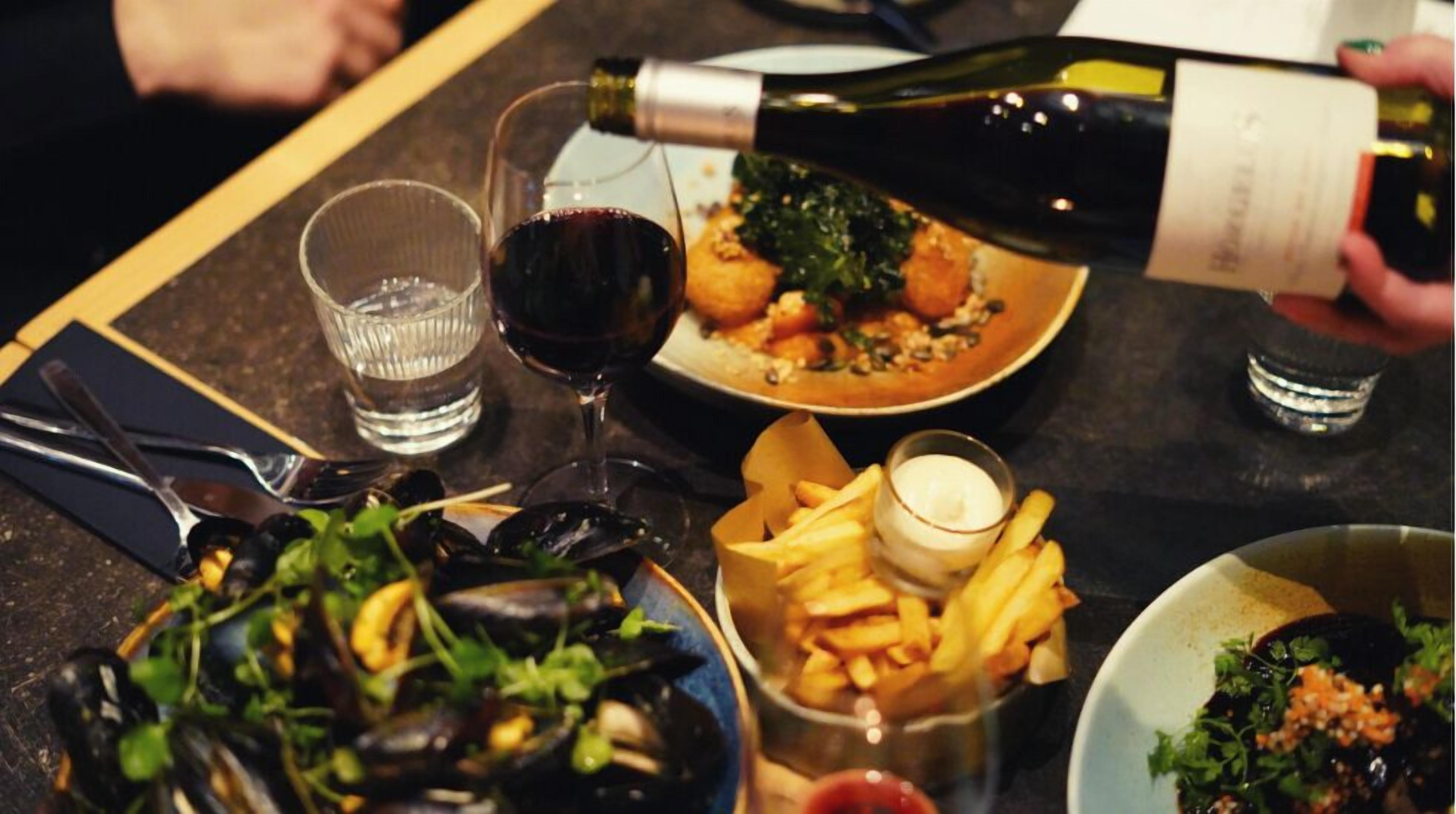 Una mesa de comedor con mejillones, papas fritas y una botella de vino siendo servida.