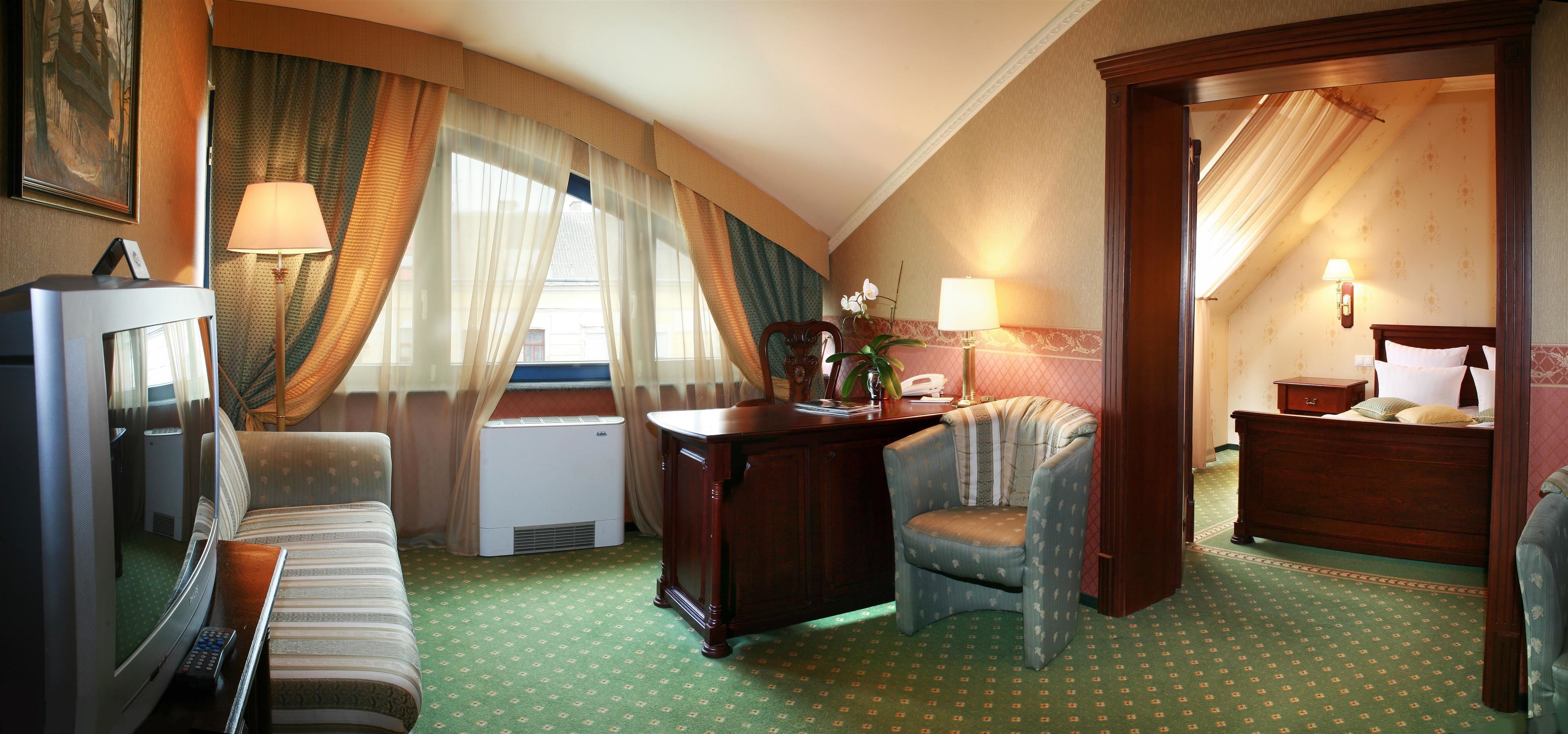Quarto de hotel com teto curvo, carpete verde estampado, sofá, escrivaninha com luminária, cadeira, cama e televisão.
