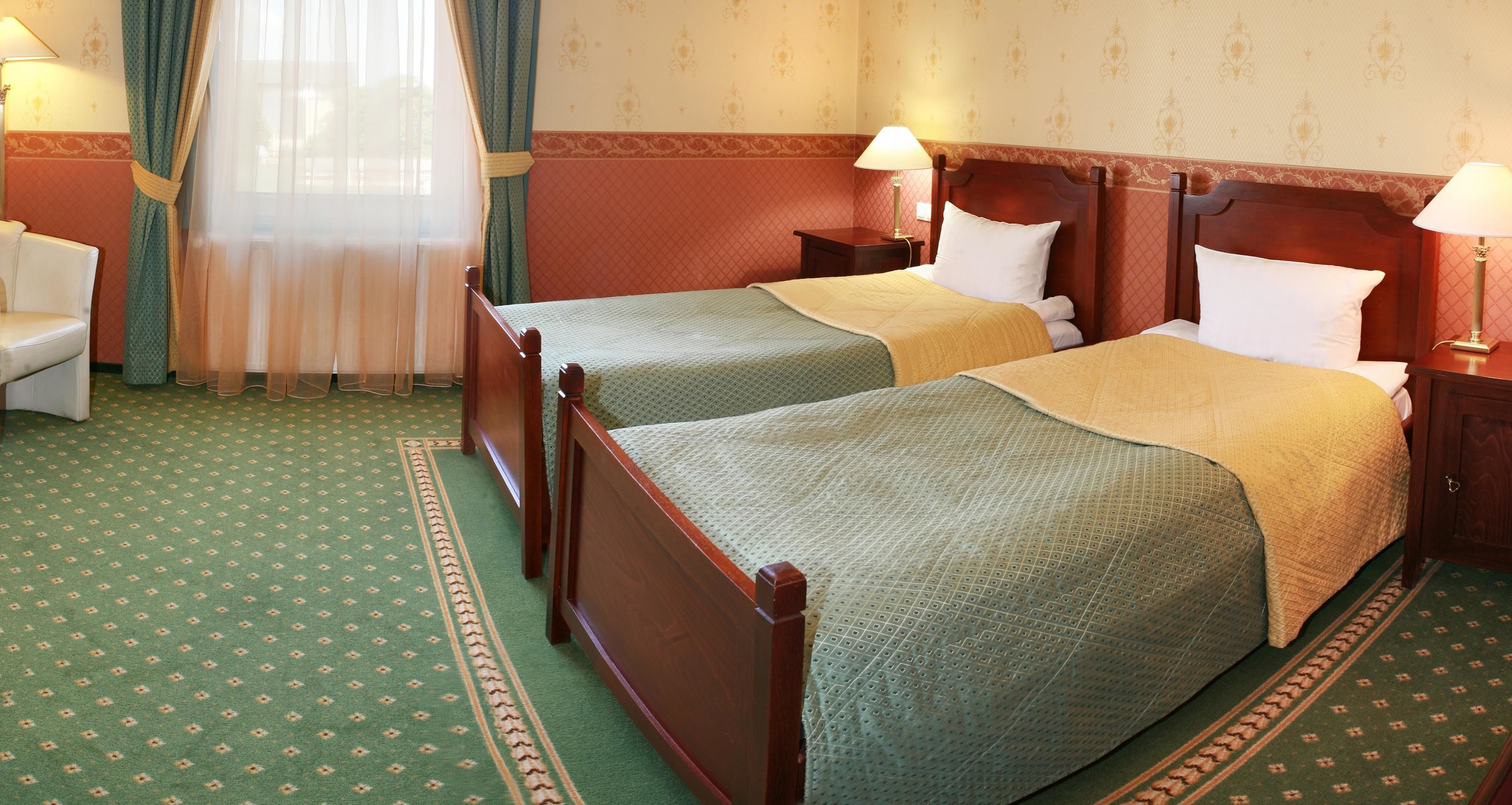 A hotel room with two wooden beds, a green carpet, and a window with sheer curtains.