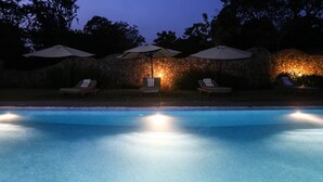 Piscina con sillas de descanso y sombrillas, rodeada de muros de piedra y árboles.