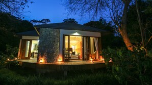 Un refugio moderno y aislado, con fachada de piedra, grandes puertas de cristal que dan a un interior bien iluminado y una terraza de madera con iluminación ambiental.