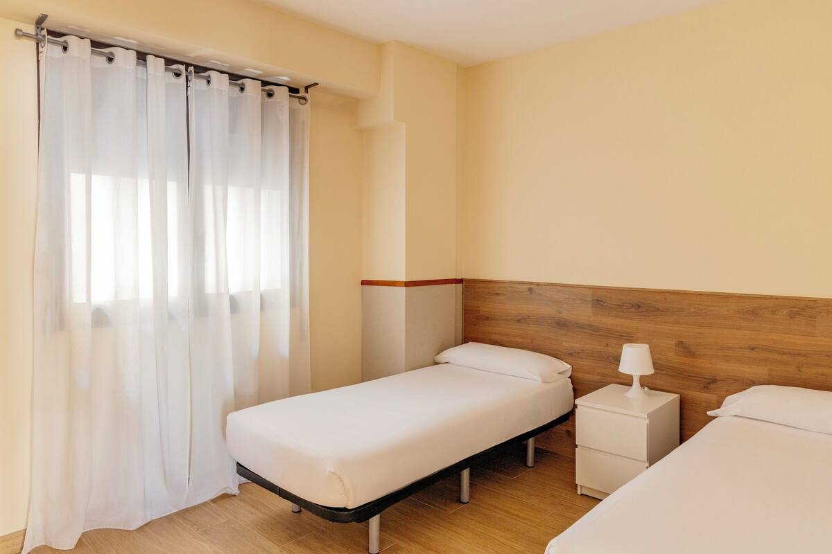 A single bed with a wooden headboard, a bedside table with a lamp, and a curtain on a window.