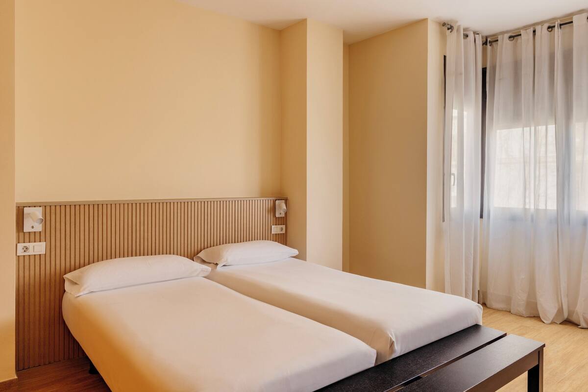 A hotel room with two beds, a wooden headboard, a bedside table, a window with sheer curtains, and a light switch on the wall.