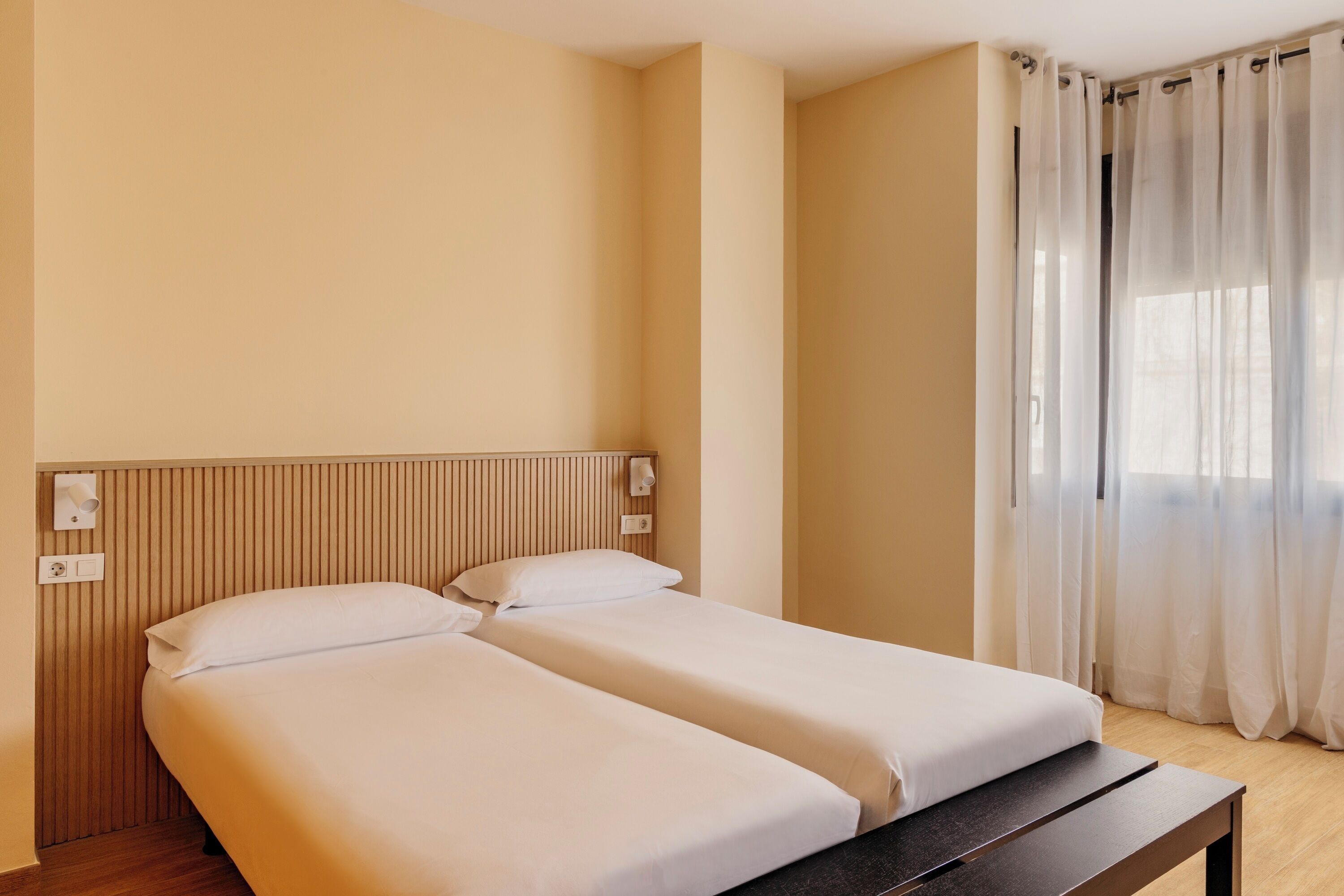A hotel room with two beds, a wooden headboard, a bedside table, a window with sheer curtains, and a light switch on the wall.