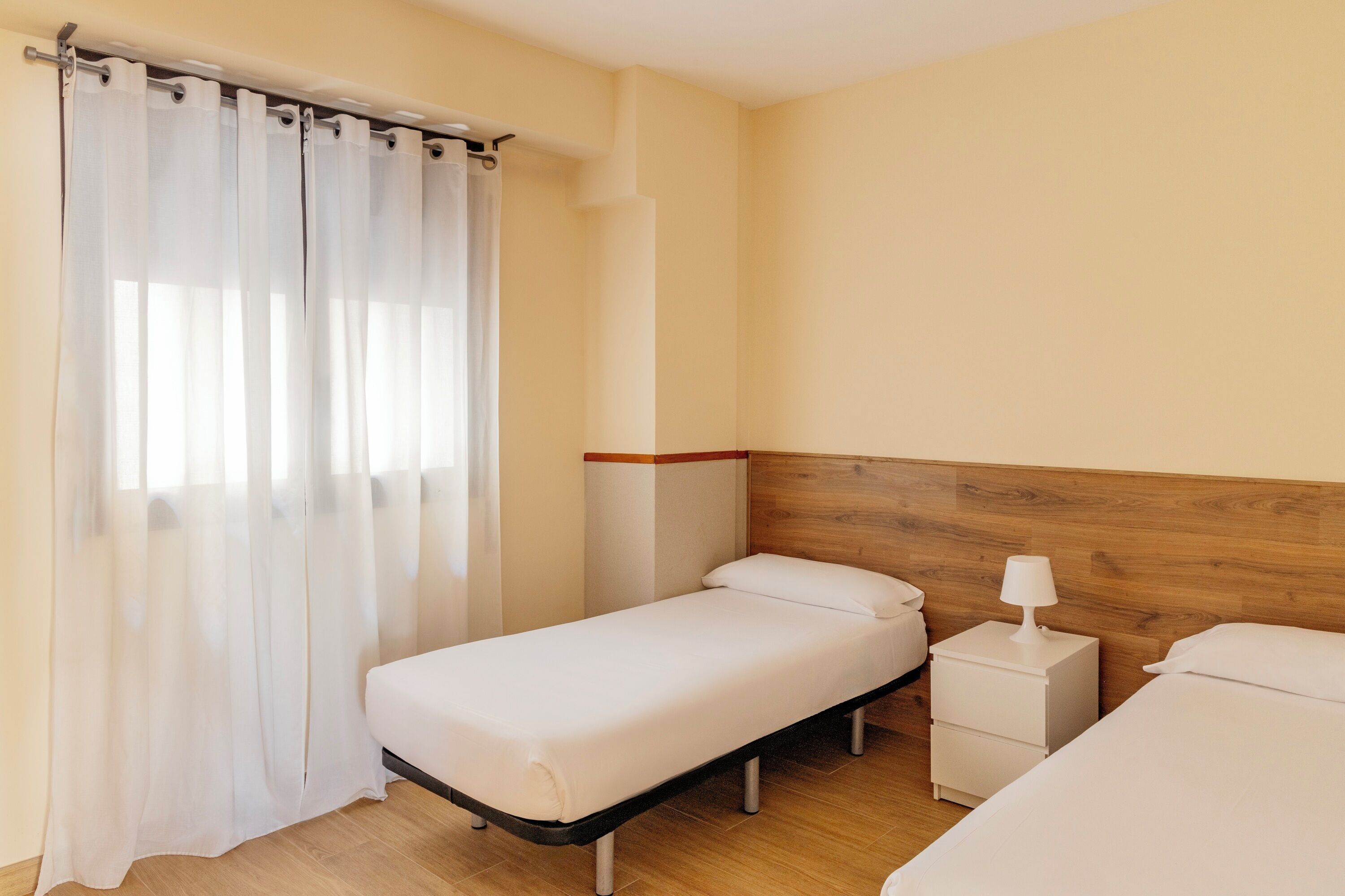 A single bed with a wooden headboard, a bedside table with a lamp, and a curtain on a window.