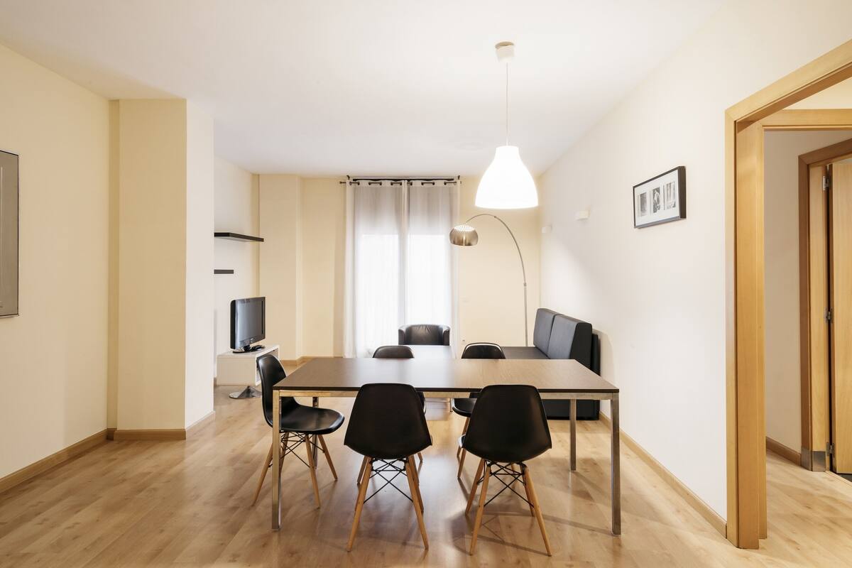 A modern dining area with a long table, four black chairs, a television, and a large pendant light.