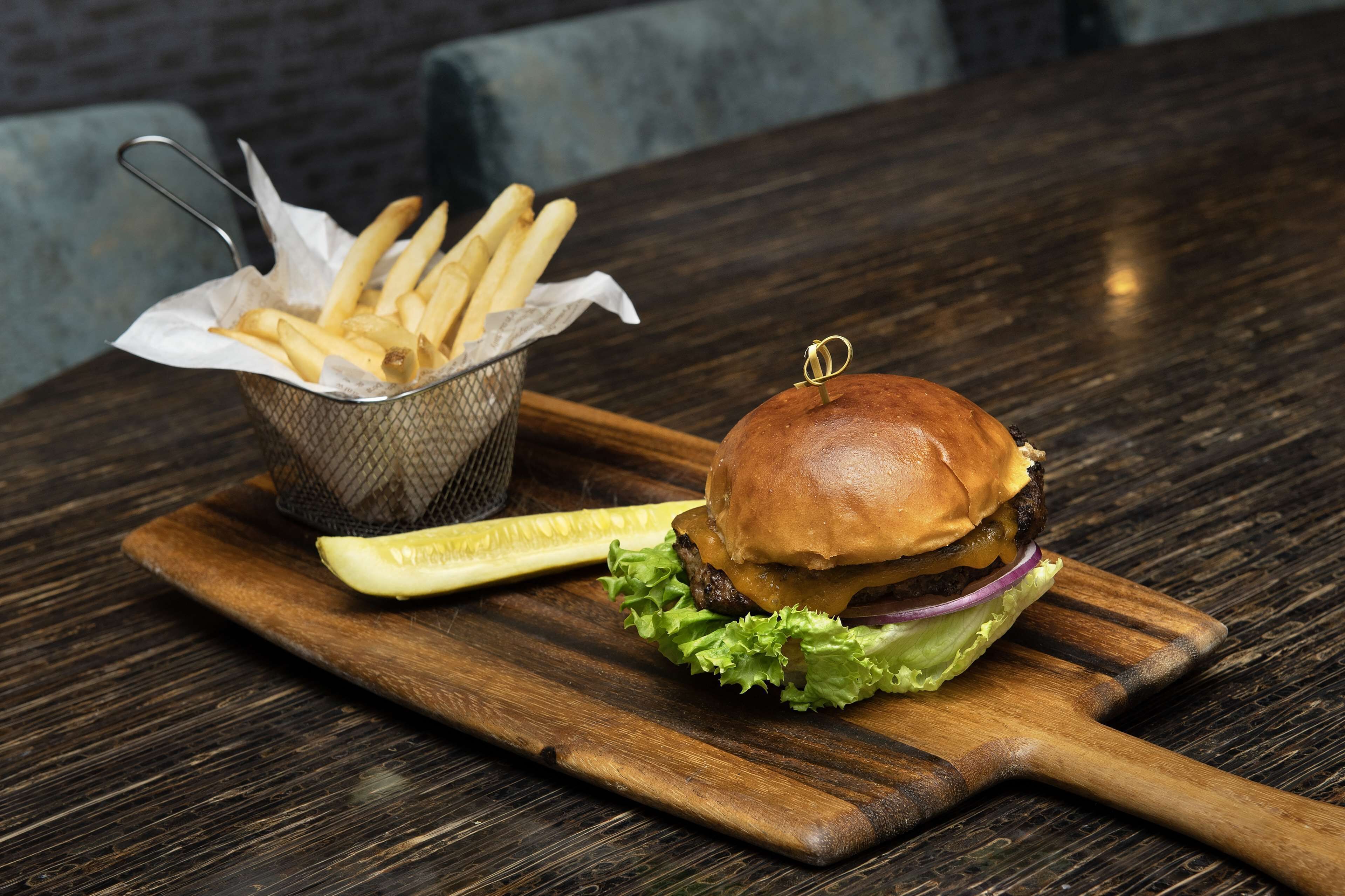 Hamburguesa con lechuga y pepinillo sobre una tabla de madera, acompañada de una cesta de papas fritas y otro pepinillo en una mesa de madera oscura.