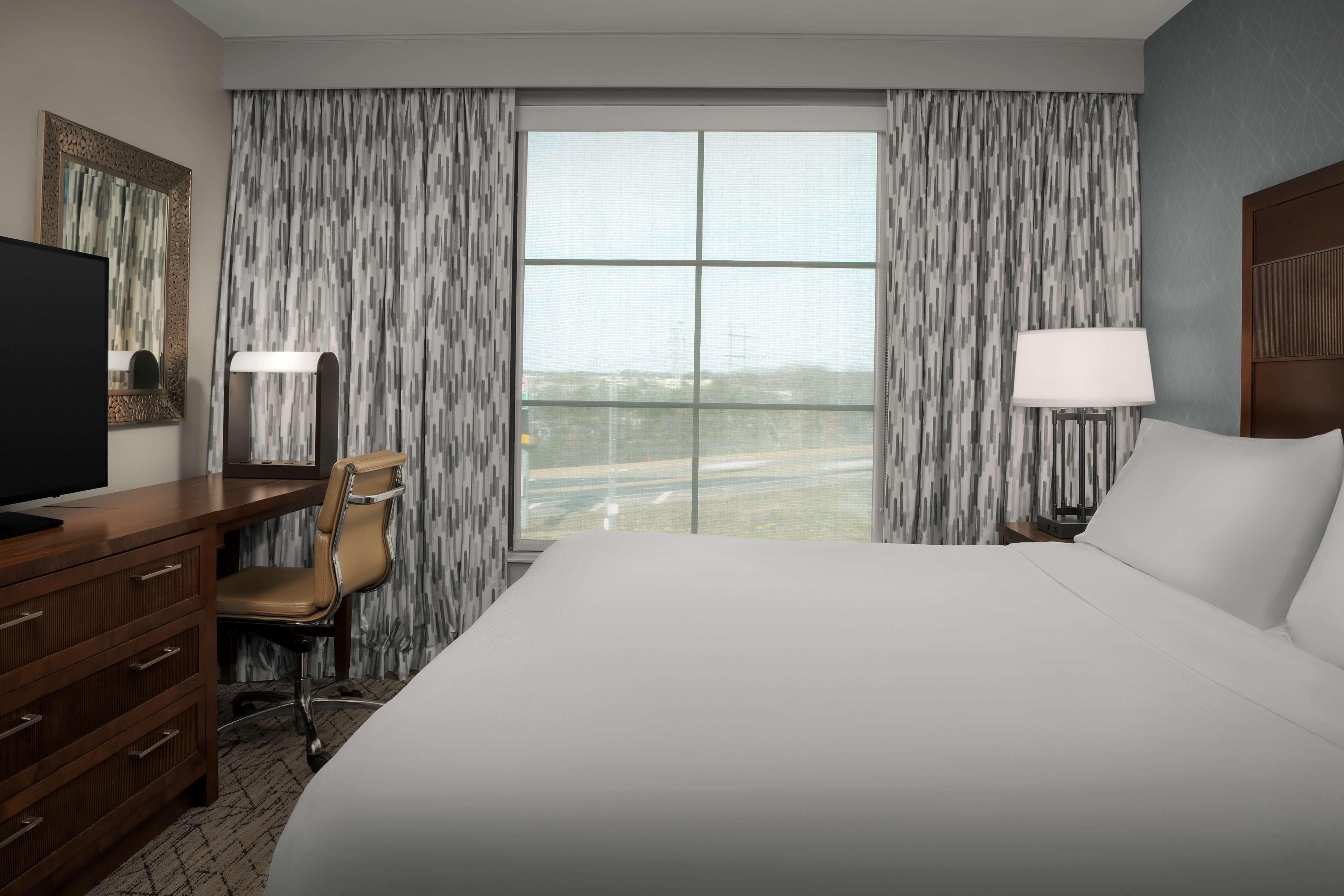 A hotel room with a large bed, a desk with a chair, a TV, and a window with curtains.
