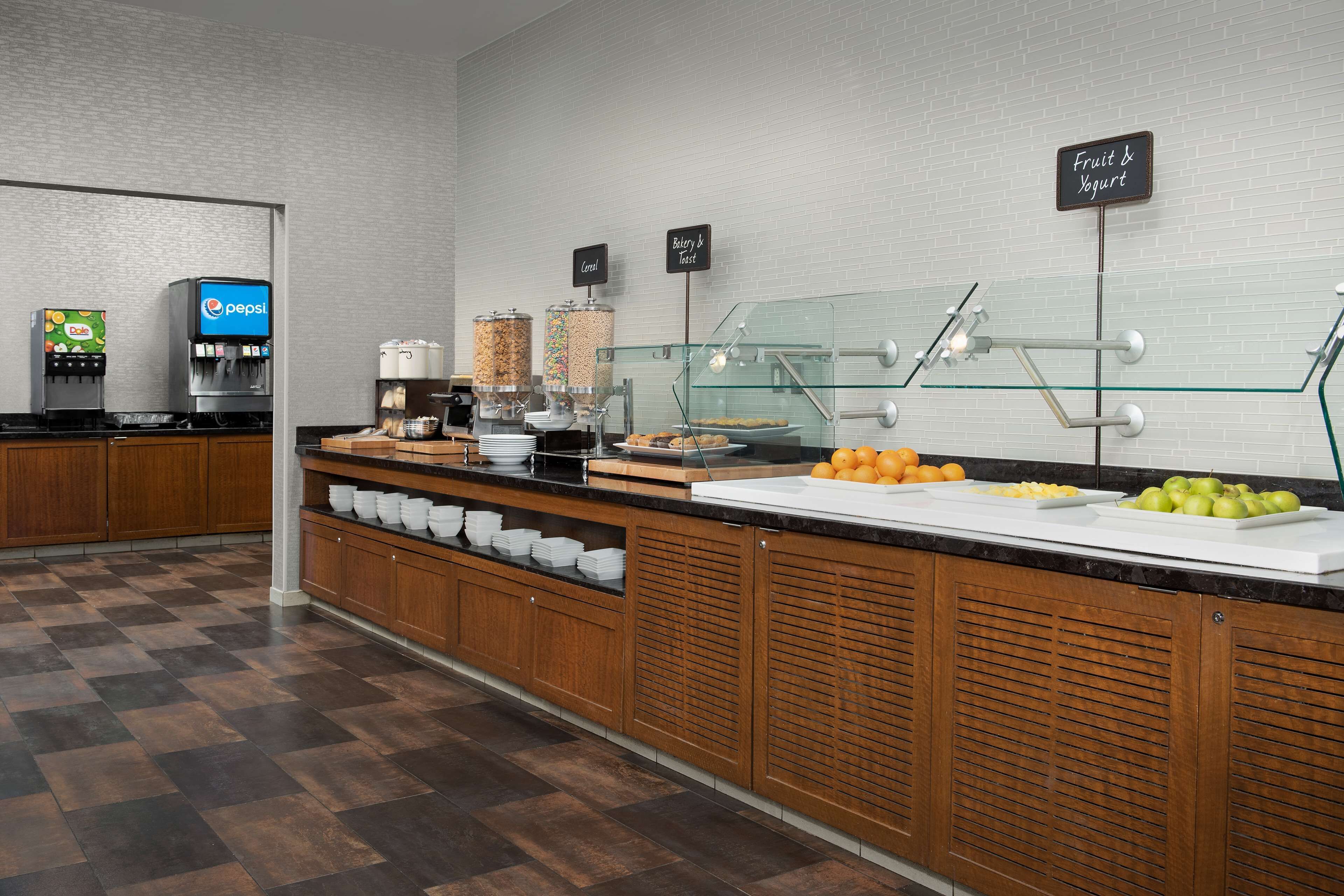 Uma área de buffet com dispensador de Pepsi, estação de frutas e iogurte e diversos alimentos expostos no balcão.