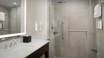 Une salle de bain moderne avec un grand miroir, un lavabo à deux robinets, une douche en verre avec une barre d’appui et des articles de toilette fixés au mur.