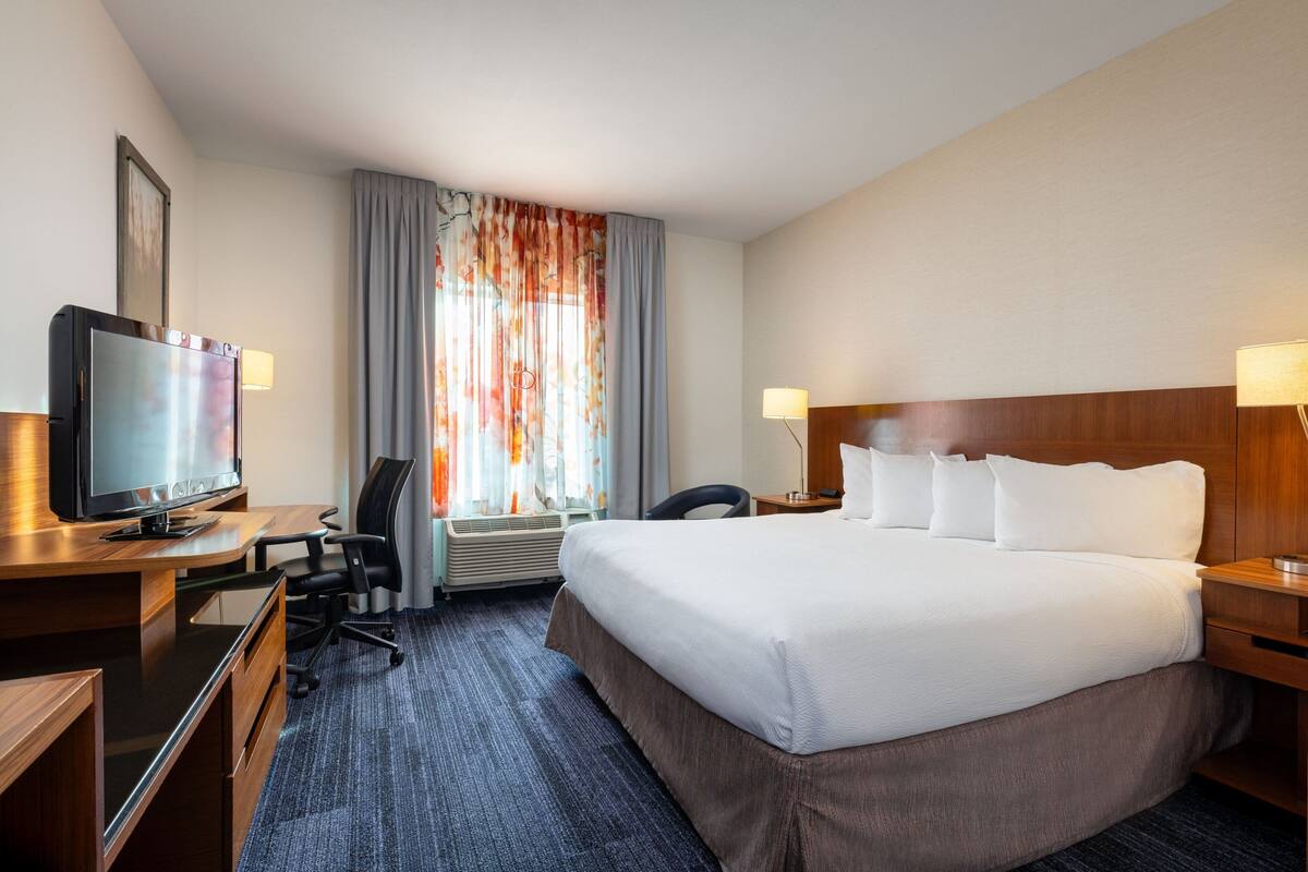 Une chambre d’hôtel avec un grand lit, un bureau avec une télévision, une chaise, une lampe de bureau et une fenêtre avec des rideaux.