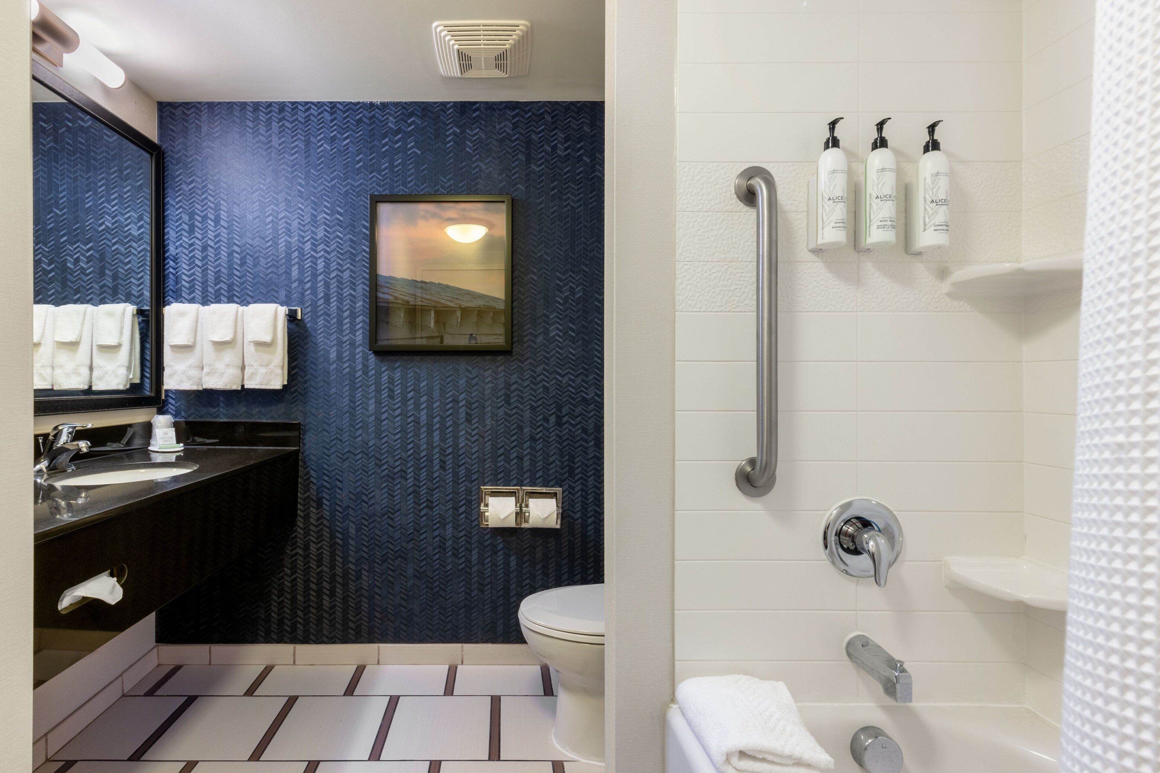 A modern bathroom with a blue patterned wall, a large mirror, a sink, a toilet, and a shower area with handrails and toiletries.