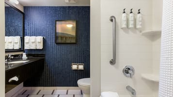 A modern bathroom with a blue patterned wall, a large mirror, a sink, a toilet, and a shower area with handrails and toiletries.