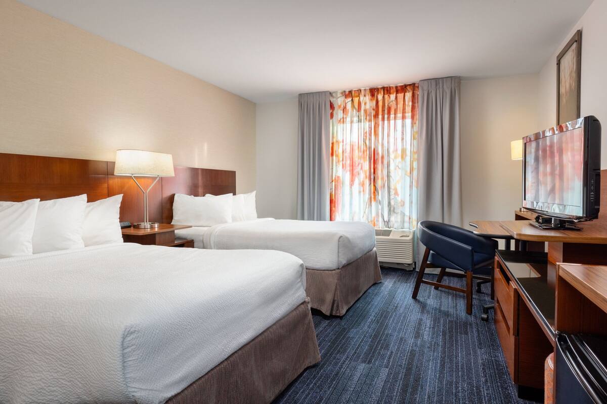 Une chambre d’hôtel avec deux lits, un bureau avec une télévision, une lampe et une fenêtre avec des rideaux.