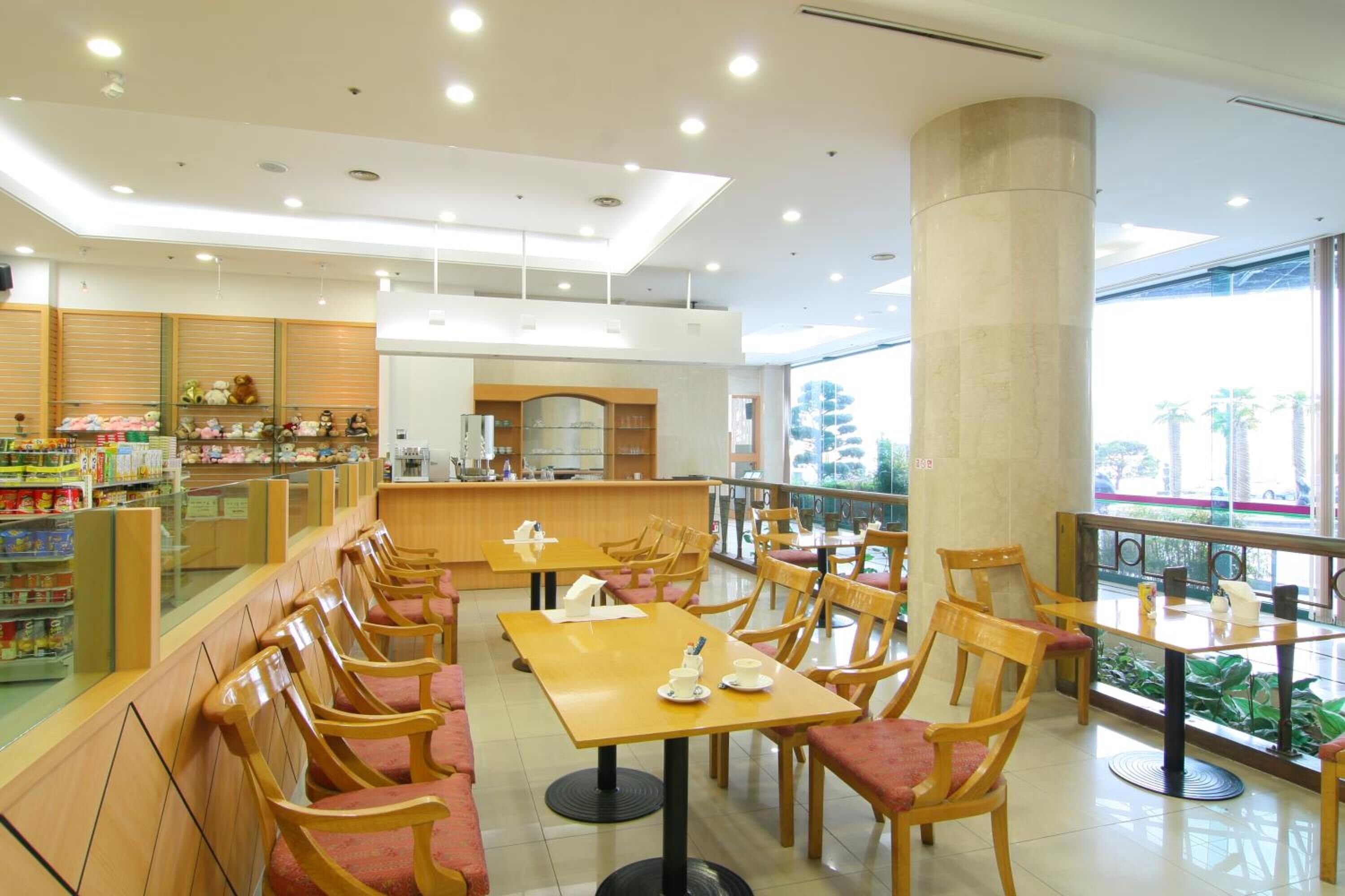A spacious cafe with wooden furniture, a counter with various items, and a view of palm trees through large windows.
