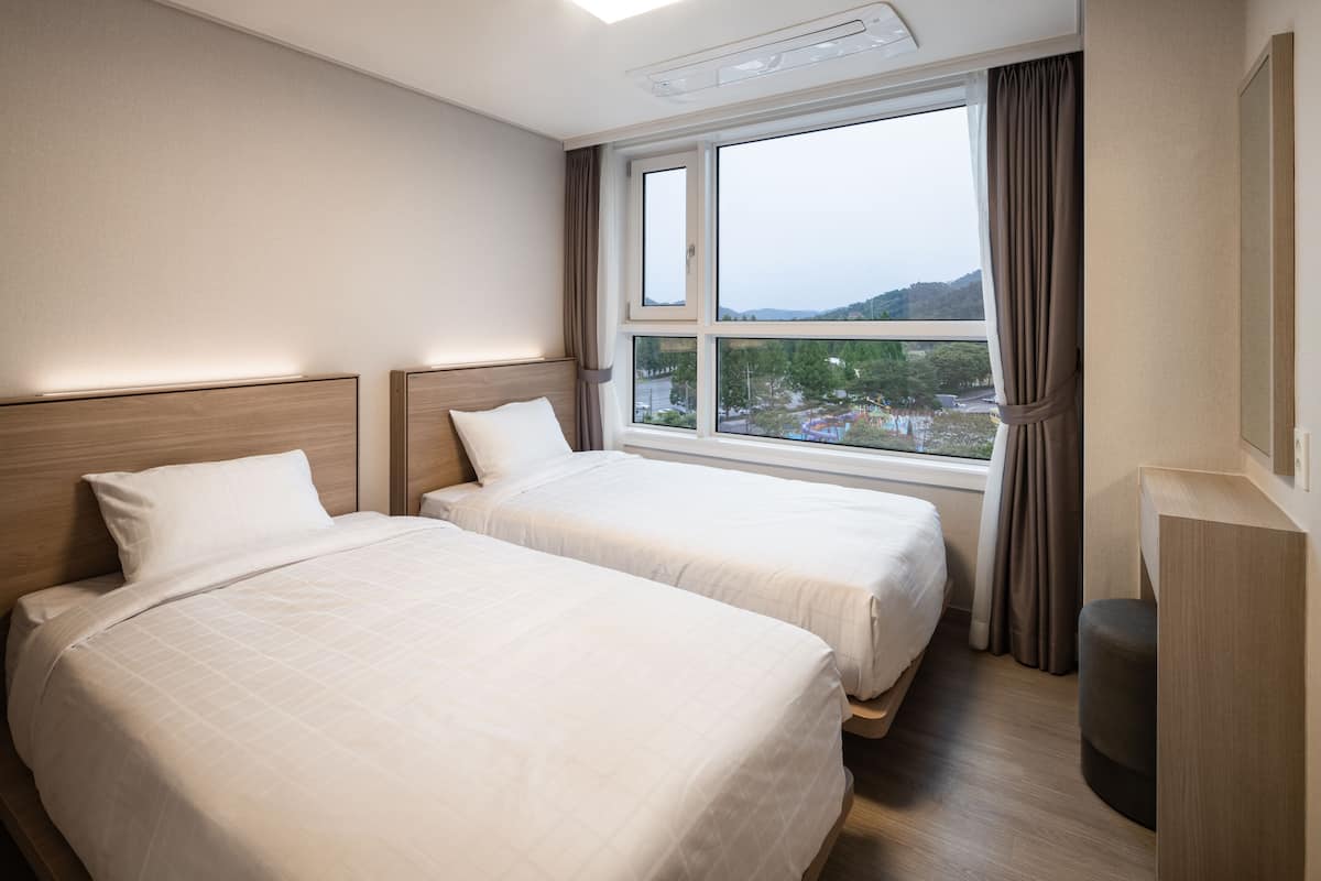 Habitación de hotel con dos camas, un ventanal con cortinas y vista a un paisaje urbano.