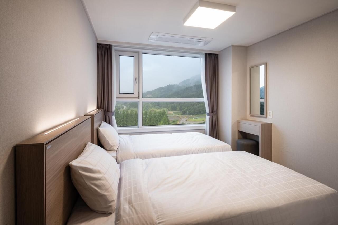 A hotel room with two beds, a large window with curtains, a bedside table, and a mirror.