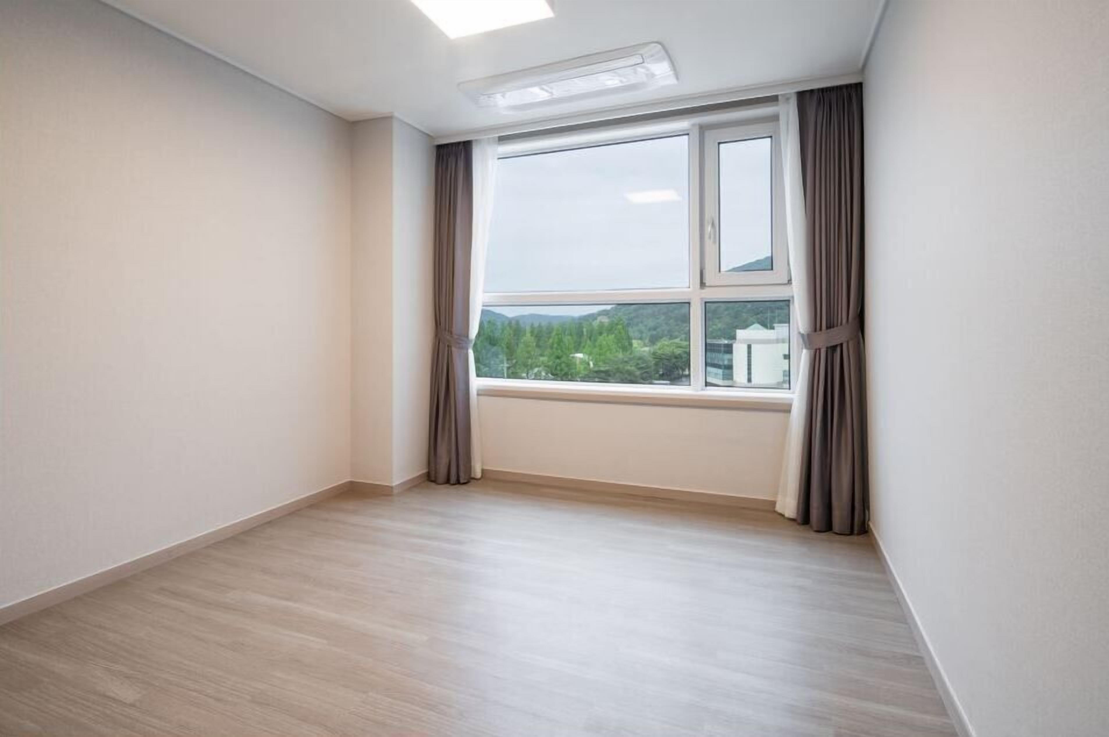 A room with a large window, light wooden flooring, and a view of greenery and buildings outside.