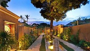 A well-lit garden pathway leading to a building with a large tree and a small water feature. - New Pondok Sara Villas (Seminyak)