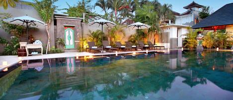 A swimming pool with lounge chairs and umbrellas, surrounded by tropical plants and a traditional building.