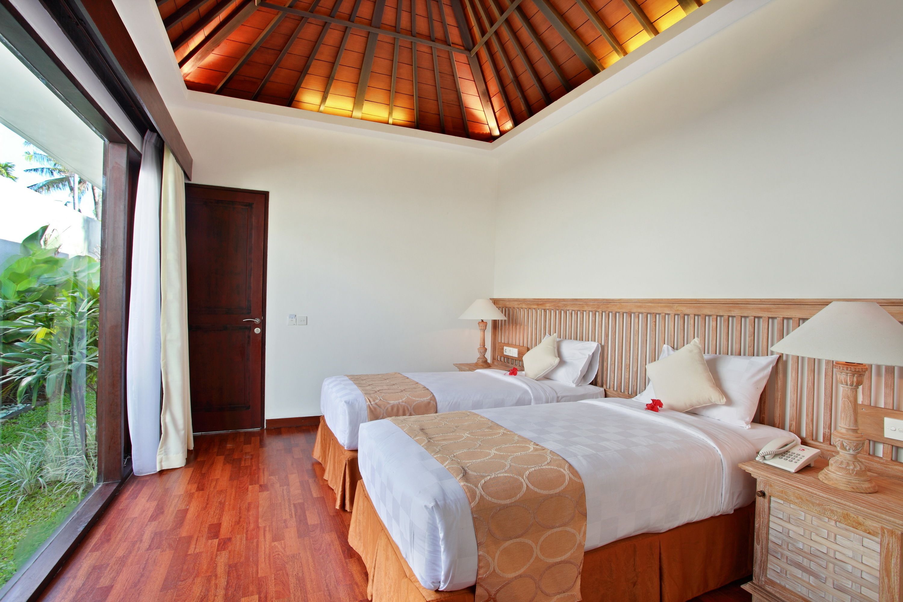 A hotel room with two beds, wooden headboards, a wooden floor, a large window, and a view of greenery outside.