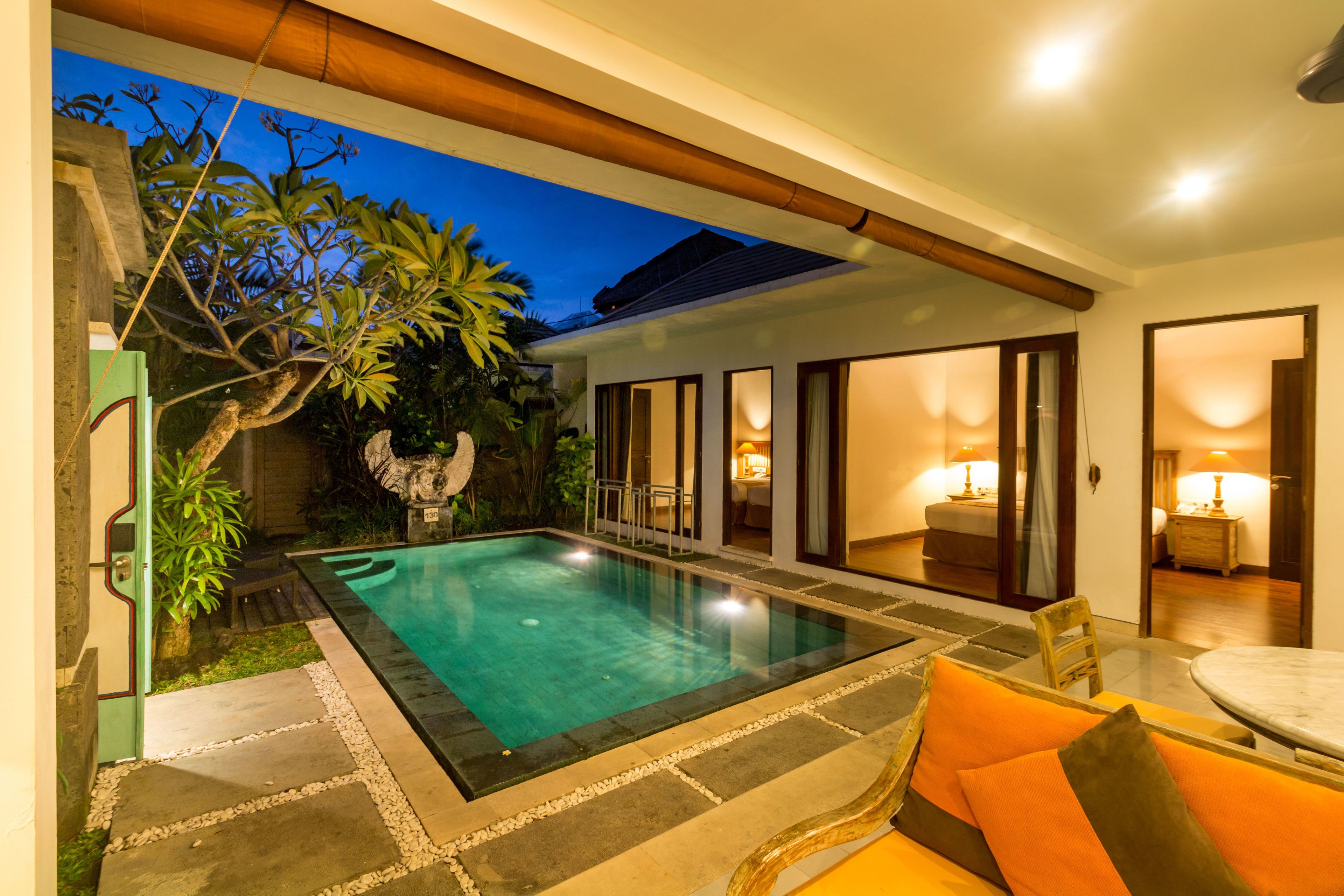 A swimming pool with a stone border, surrounded by a patio area with a wooden ceiling and a view of a room with a bed and a lamp.