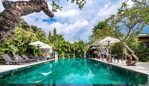 A clear blue pool with lounge chairs and umbrellas, surrounded by lush greenery and a large tree trunk. - New Pondok Sara Villas (Seminyak)
