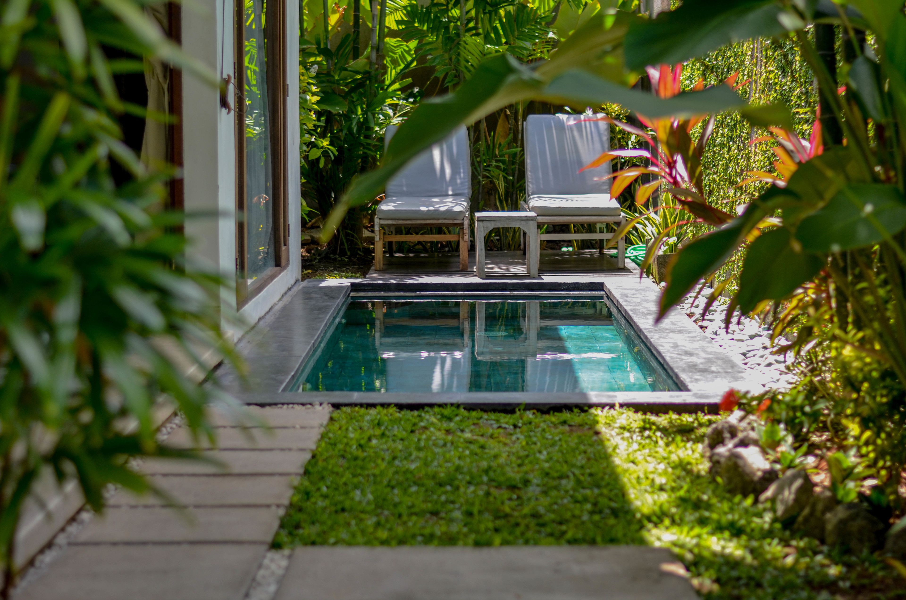 Un área de piscina con dos sillones de descanso y una mesita, rodeada de exuberante vegetación y una piscina cristalina.
