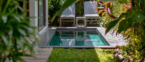 A pool area with two lounge chairs and a small table, surrounded by lush greenery and a clear pool.