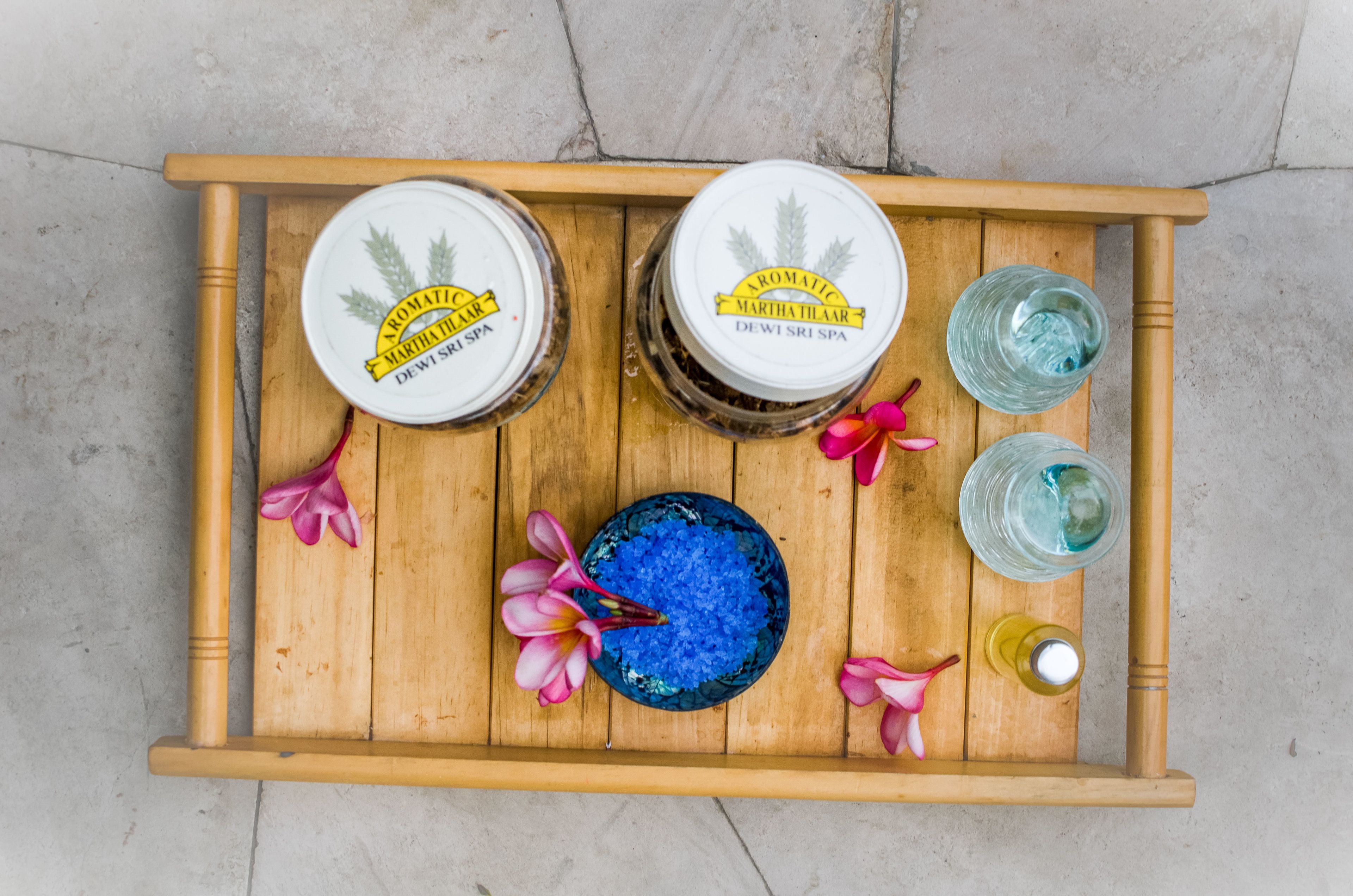 Un plateau en bois sur lequel reposent deux pots étiquetés « AROMATIC MARTHA PILAR DEWI SRI SPA », un bol bleu orné de fleurs roses et deux verres d’eau.