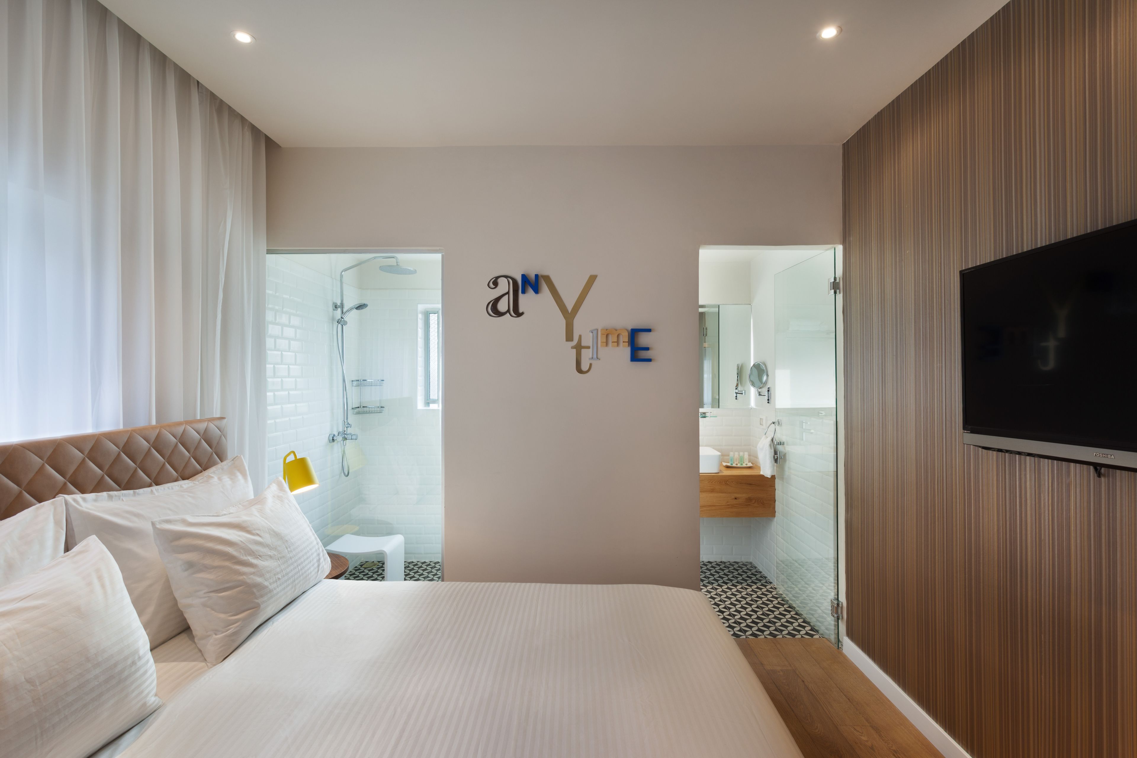 A modern hotel room with a large bed, a TV mounted on the wall, and a bathroom visible through a glass door.