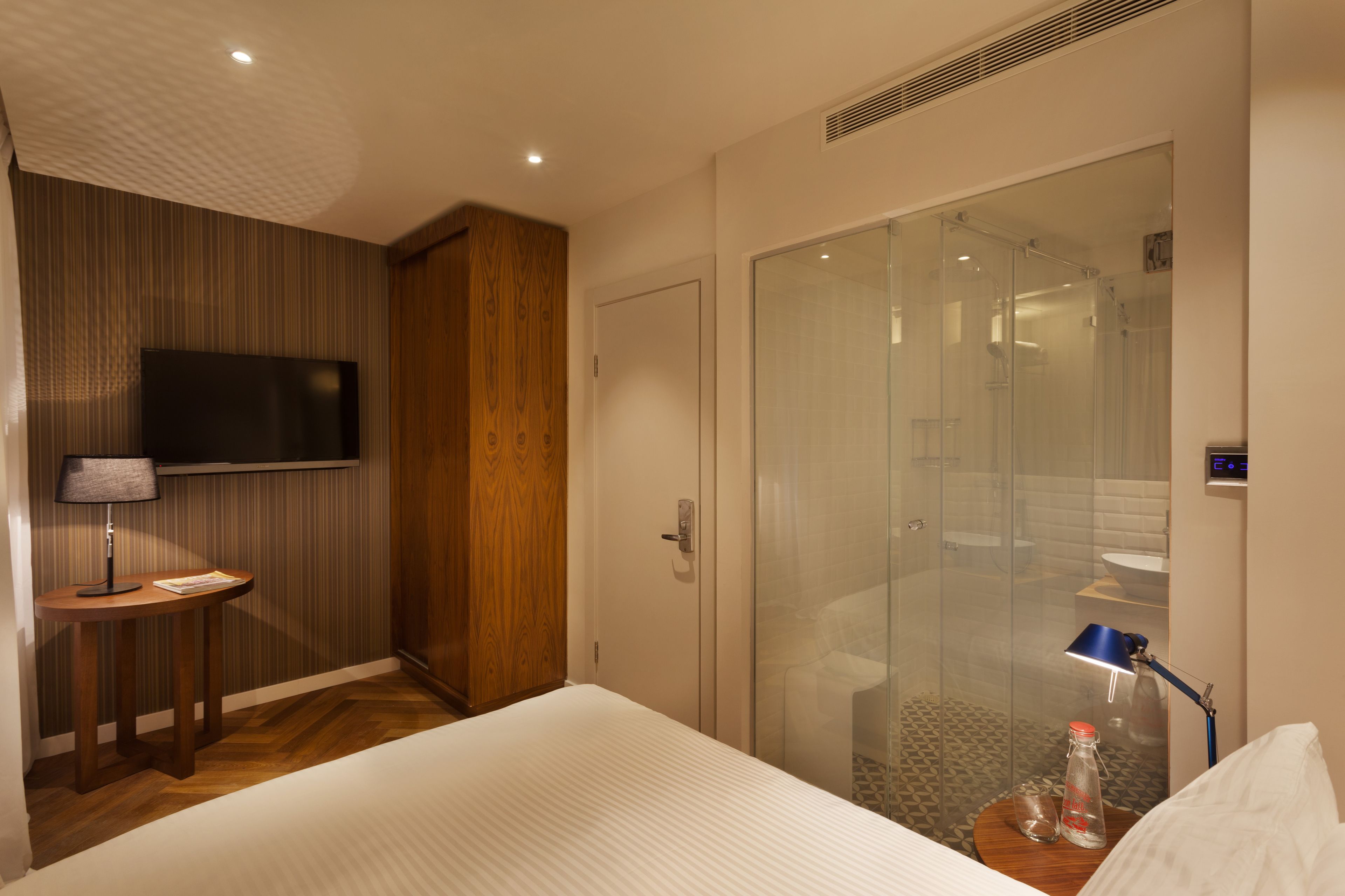 A hotel room with a bed, a TV, a glass shower, and a small table with a lamp.