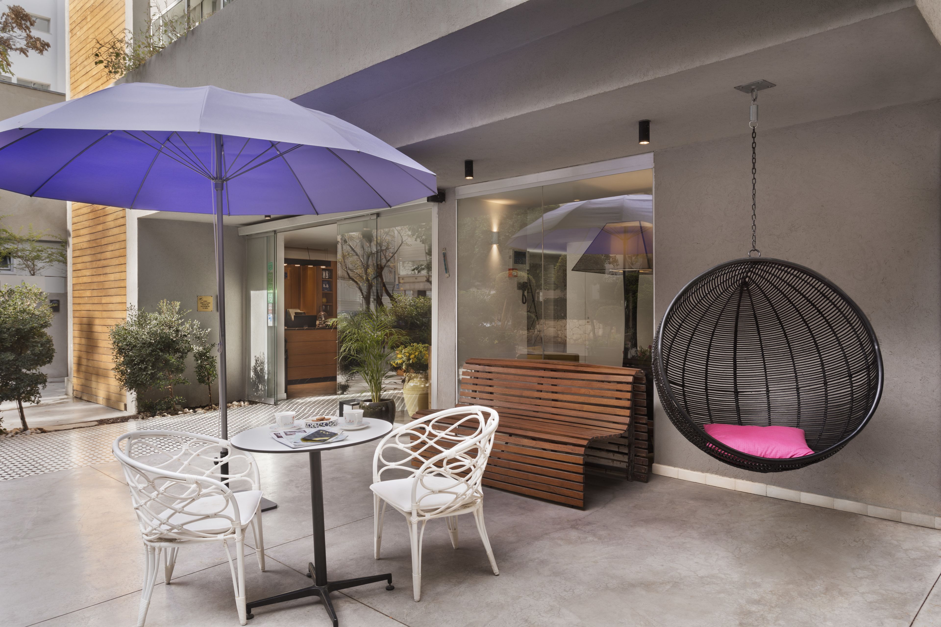Un espace extérieur aménagé avec une table ronde, deux chaises blanches, un parasol violet et un fauteuil suspendu doté d'un coussin rose.