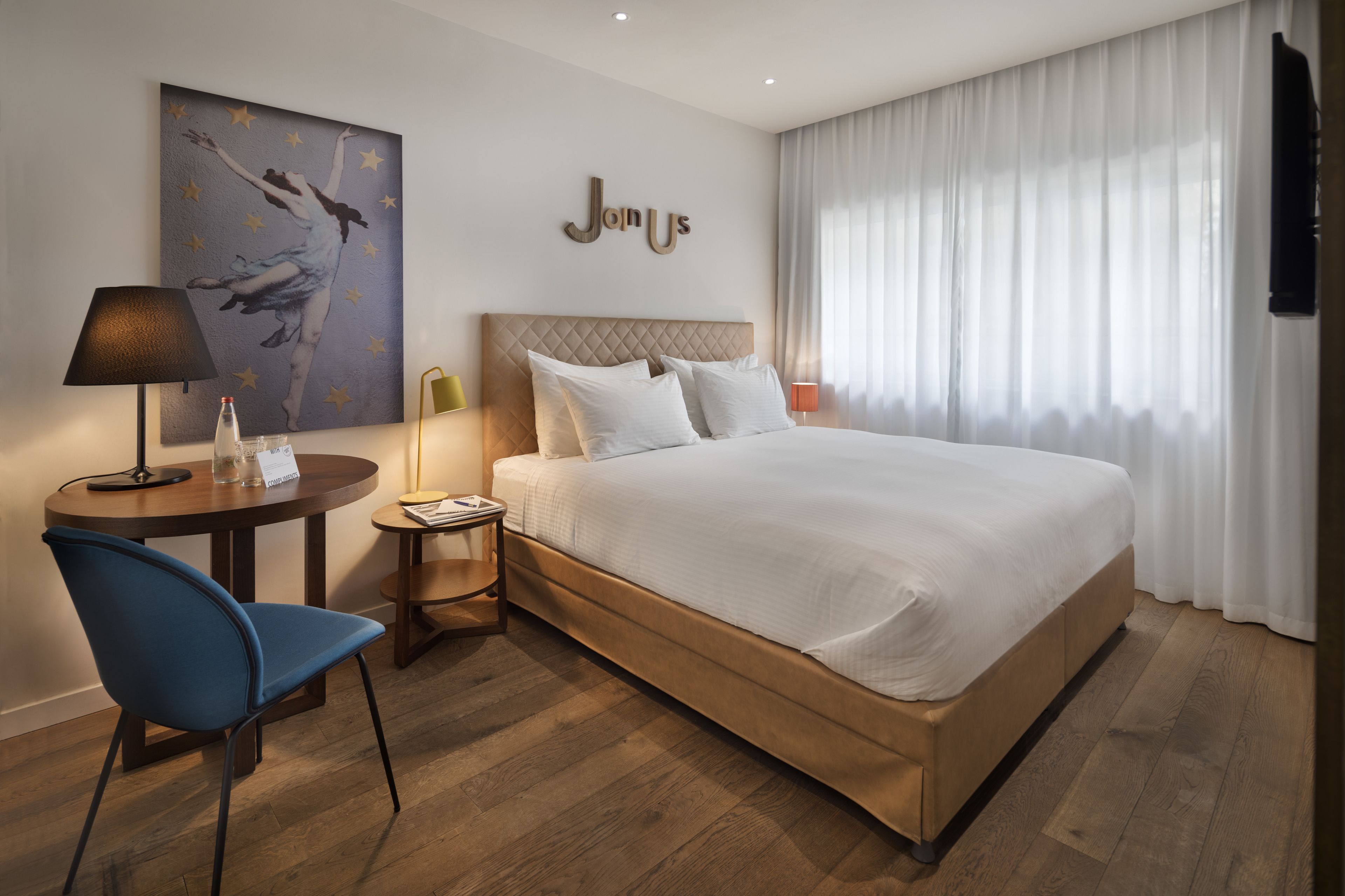 A hotel room with a large bed, a desk with a chair, a painting on the wall, and a sign that reads "Janus".