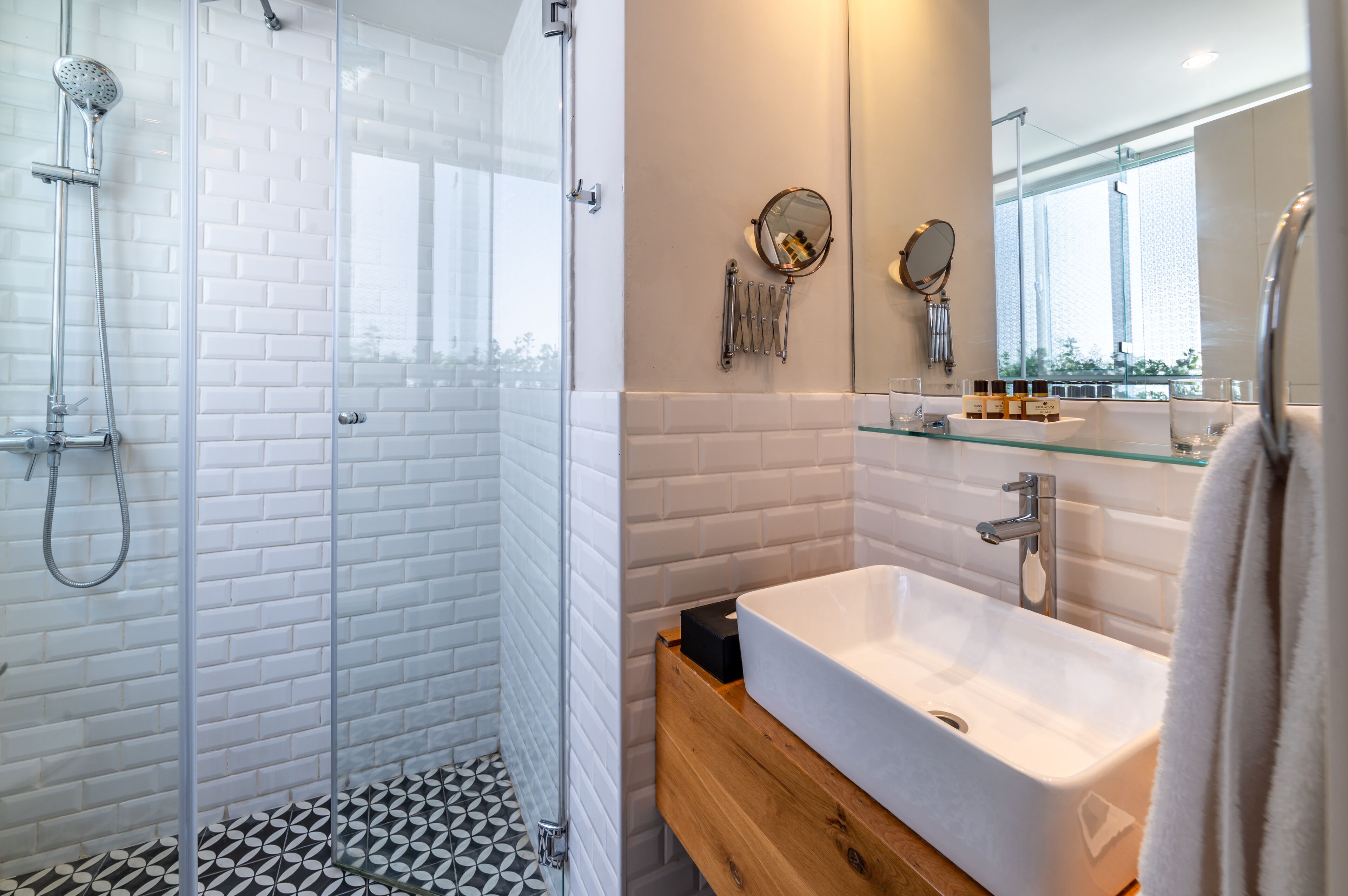 A modern bathroom with a glass-enclosed shower, white tiled walls, a wooden vanity, a white sink, and a large mirror.