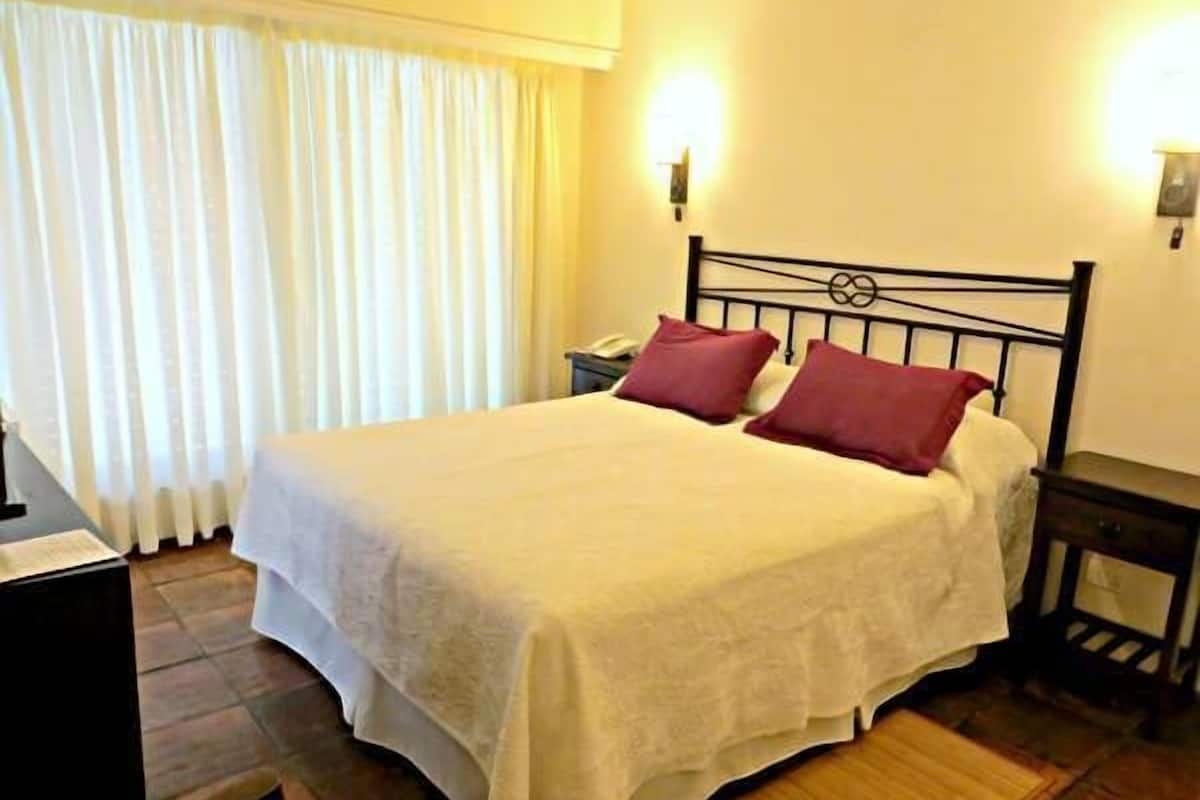 A neatly made bed with a white bedspread and two red pillows, a bedside table with a lamp, and a window with white curtains.