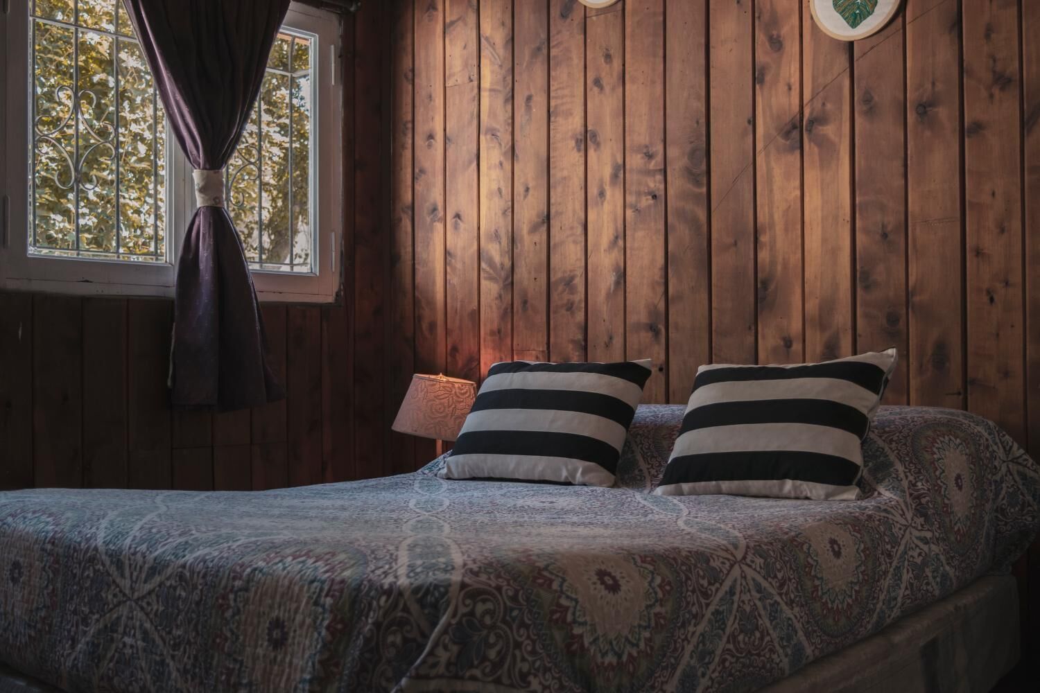 Una habitación con paneles de madera, una cama, dos almohadillas a rayas, una colcha estampada, una ventana con cortinas y un reloj de pared.