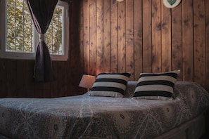 Een kamer met houten lambrisering, een bed, twee gestreepte kussens, een beddegoed met dessin, een raam met gordijnen en een wandklok.