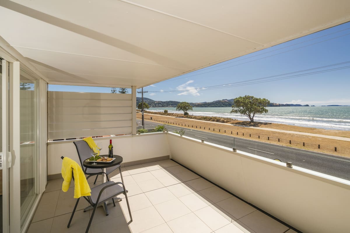A balcony with a table, chairs, and a yellow towel. It overlooks a beach and a road with trees and hills in the distance.