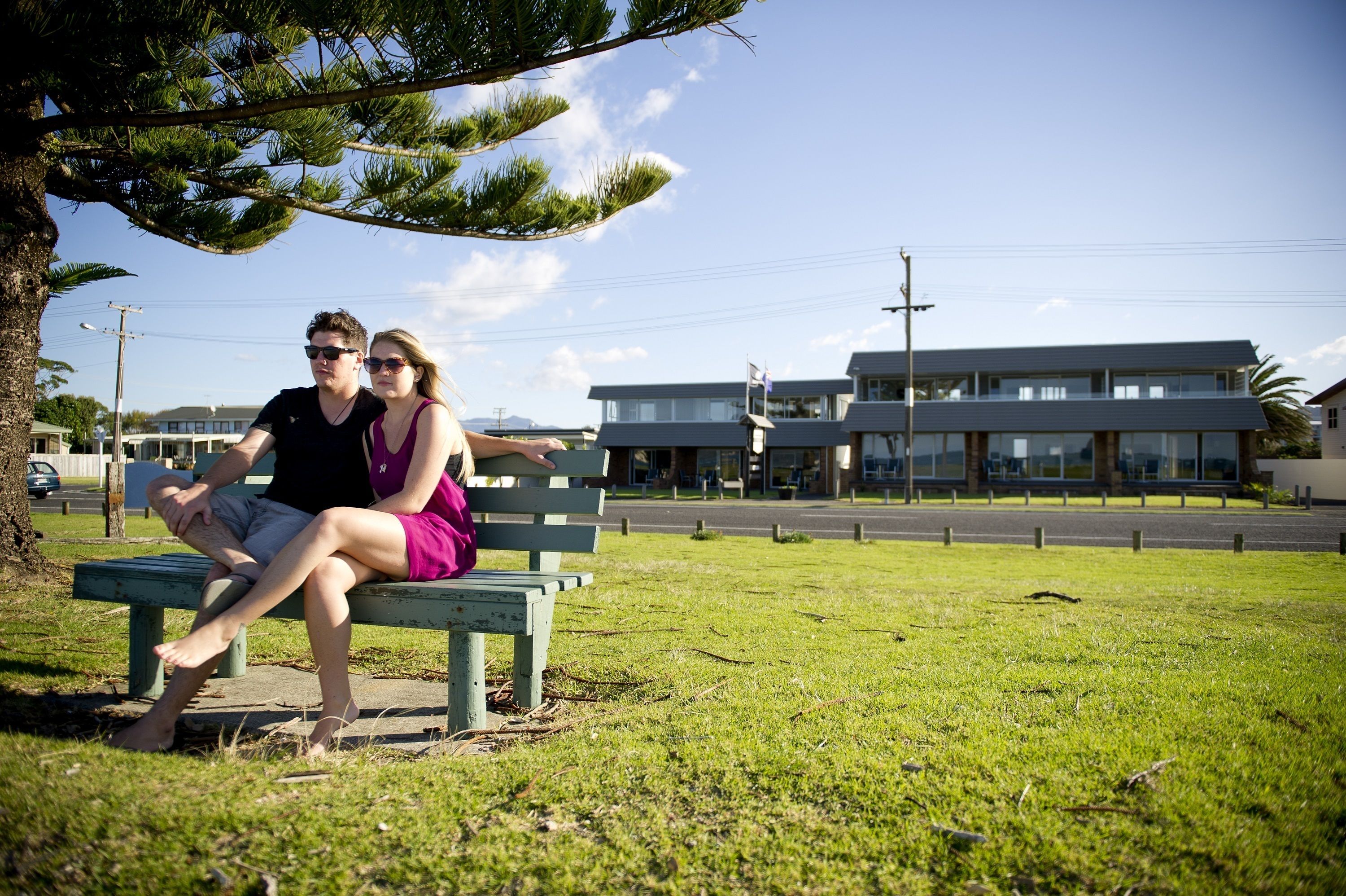 A couple sitting on a bench in a grassy area with buildings in the background.