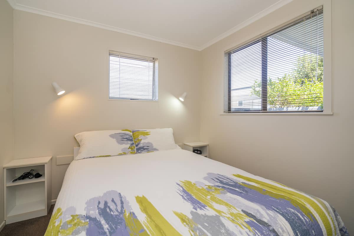 A bedroom with a bed, two bedside tables, and two windows with blinds.