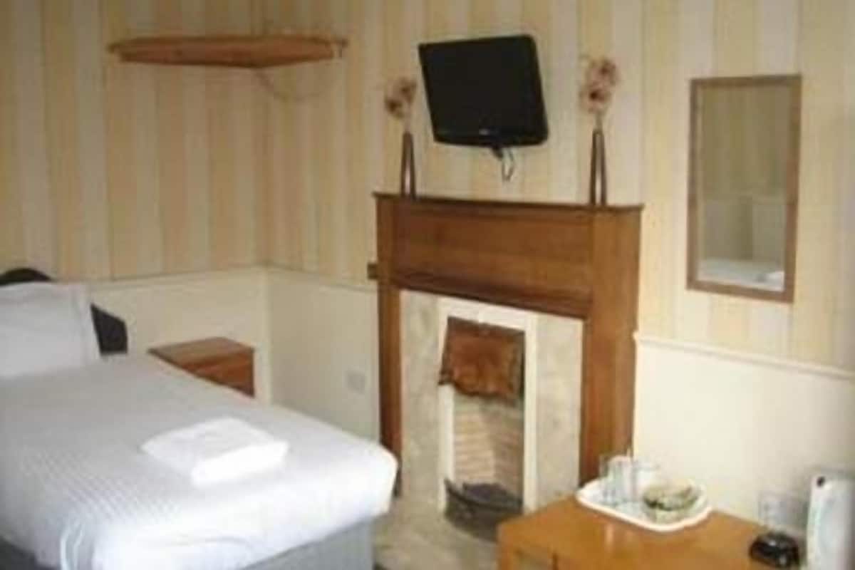 A hotel room with a bed, a wooden side table, a small table with a cup and saucer, a television mounted above a fireplace, and a mirror on the wall.