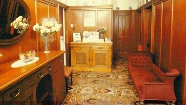 Un couloir avec des boiseries, un miroir, un vase de fleurs, un meuble en bois et une chaise rouge.