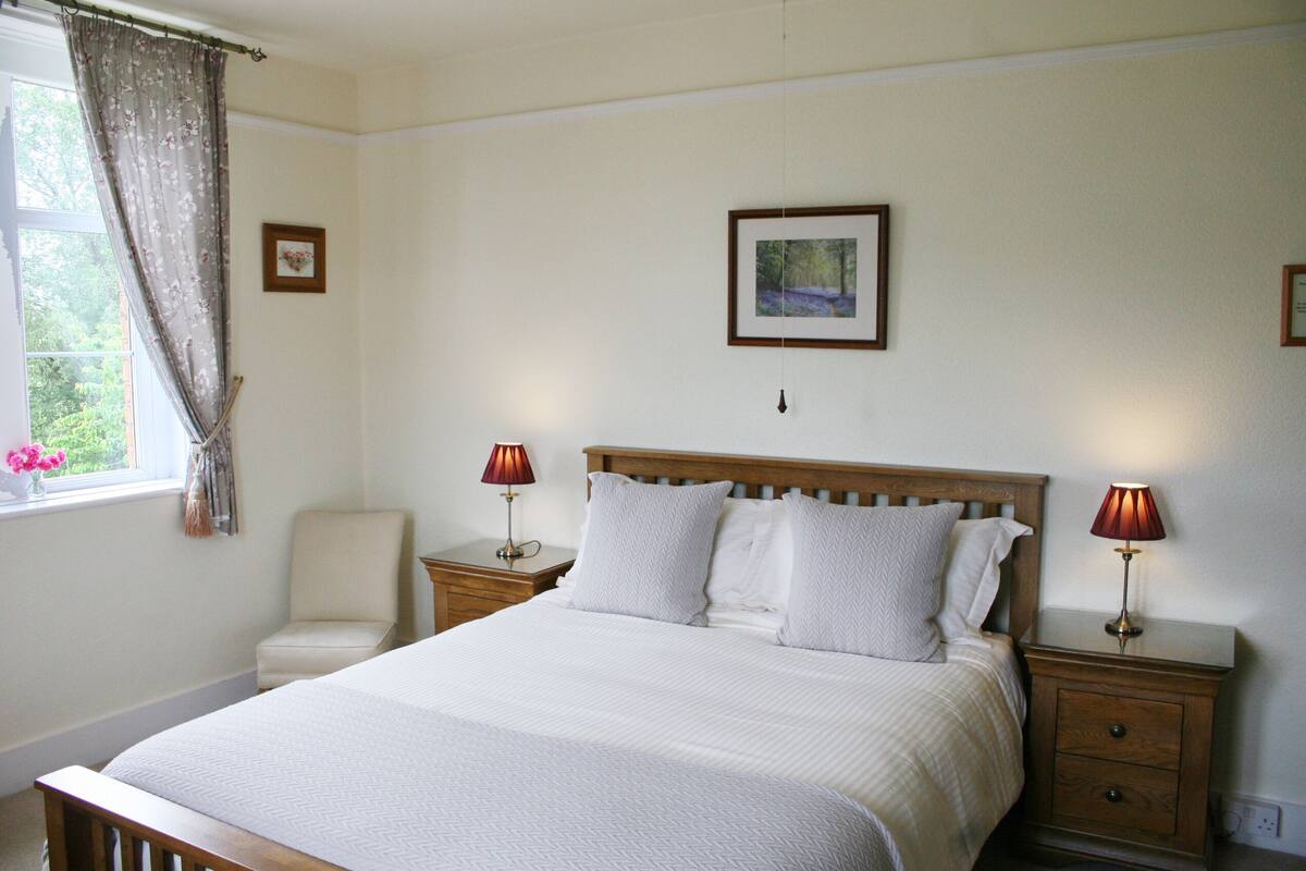 Un dormitorio con una cama grande, dos mesitas de noche con lámparas, una silla y una ventana con cortinas.