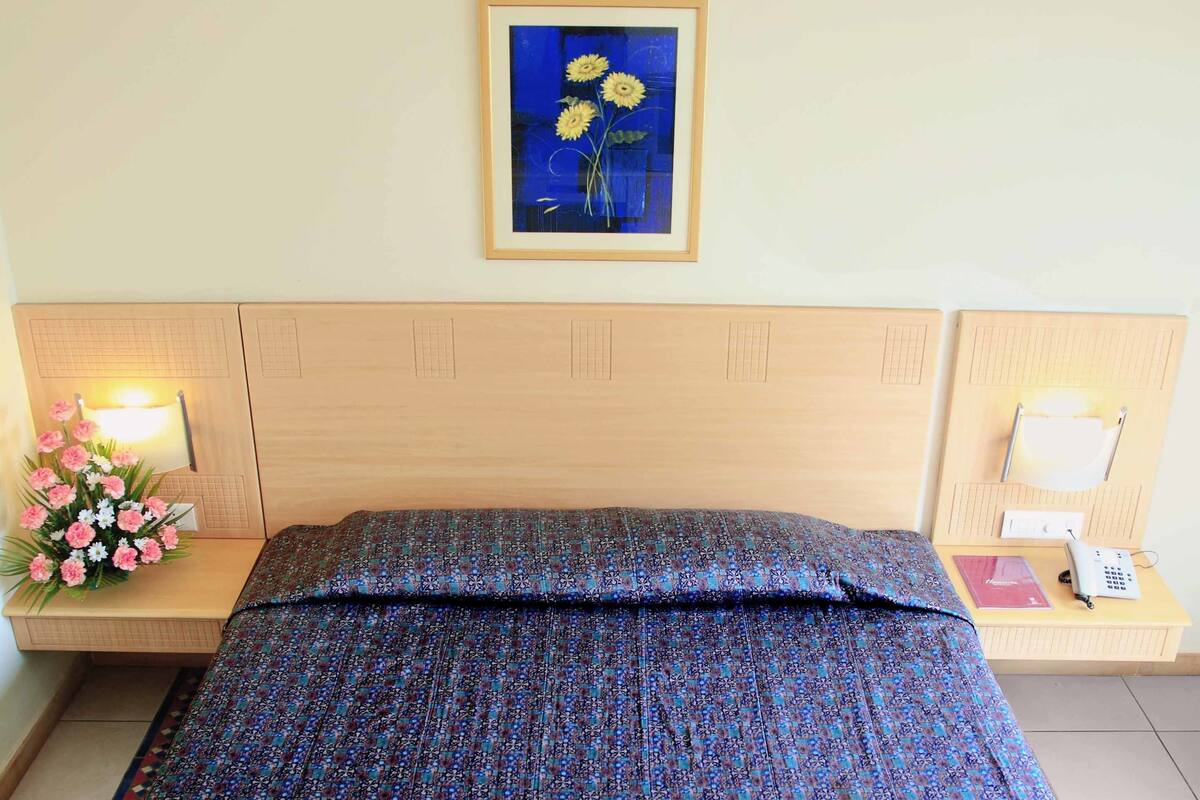 A hotel room with a bed, a nightstand with a lamp, a framed picture on the wall, and a floral arrangement on the nightstand.