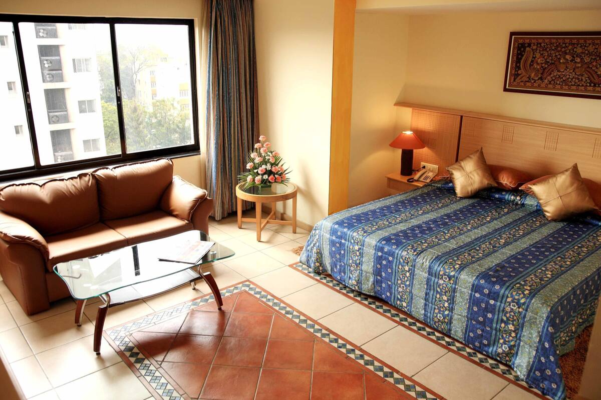 A hotel room with a bed, a sofa, a small table, and a window with a view of buildings.
